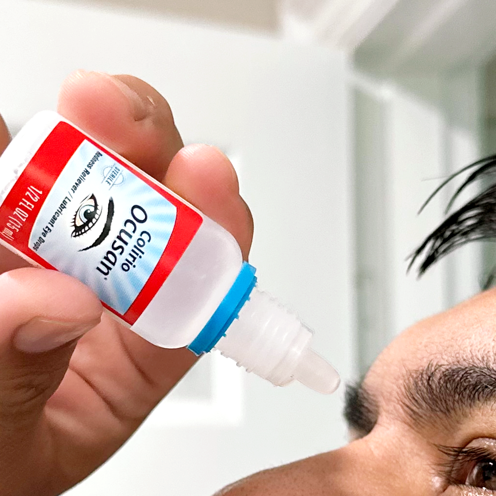 A close-up of a person holding an eye drop bottle labeled 'Colirio Ocusan' near their face, demonstrating application.