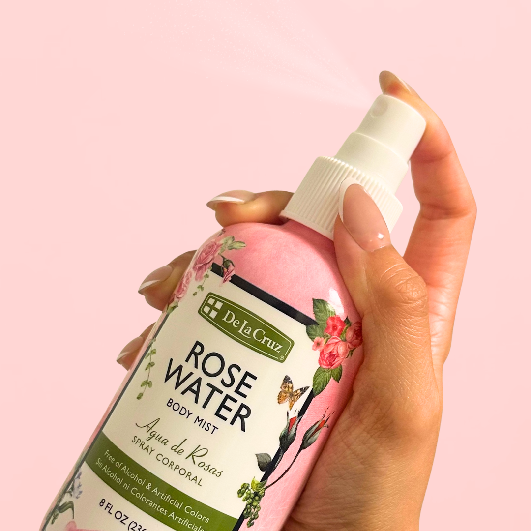 A hand holding a pink bottle of De la Cruz Rose Water Body Mist against a solid pink background.