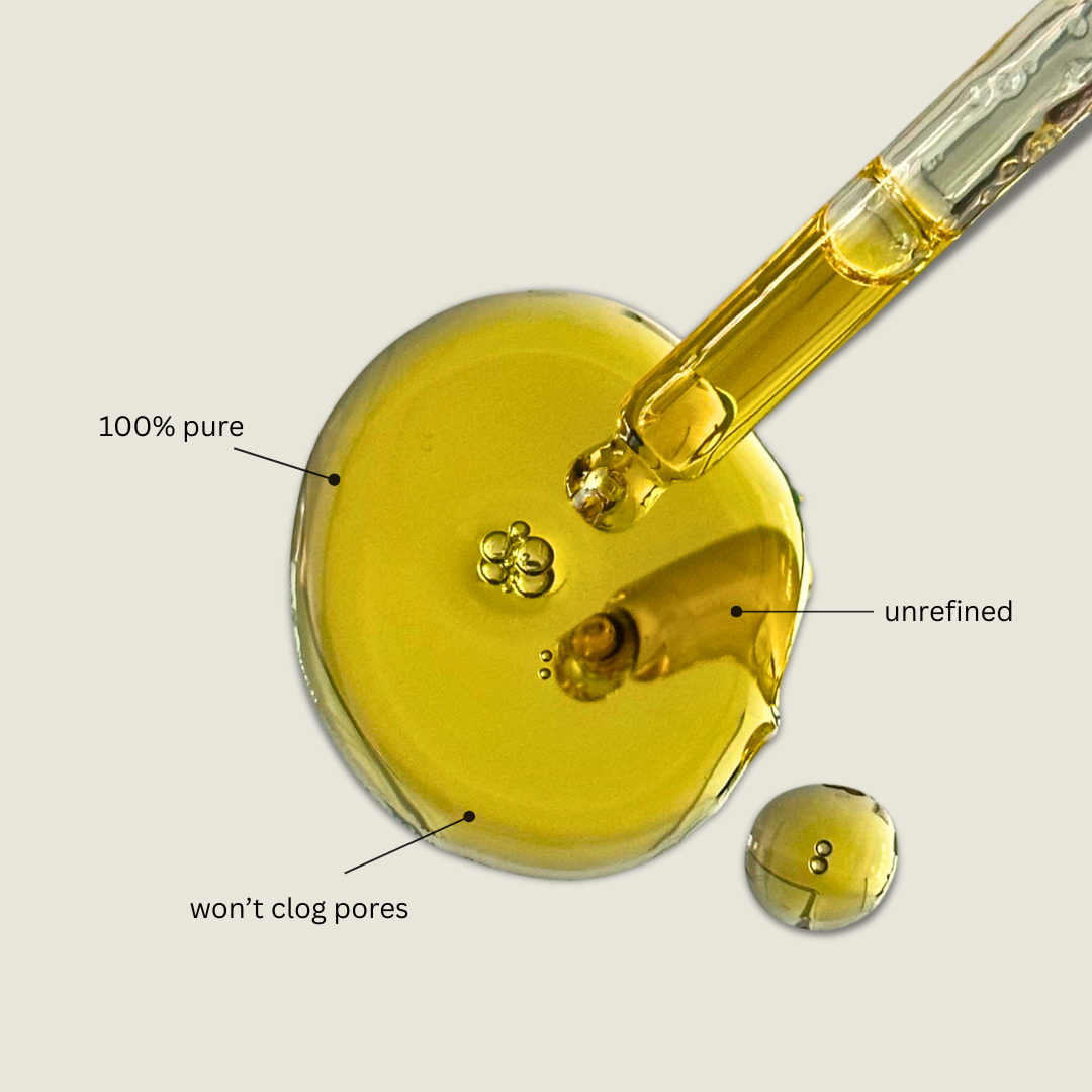A close-up of a golden-yellow cosmetic oil droplet with a glass pipette above it, plus callout labels reading '100% pure', 'unrefined', and 'won't clog pores' on a light beige background.