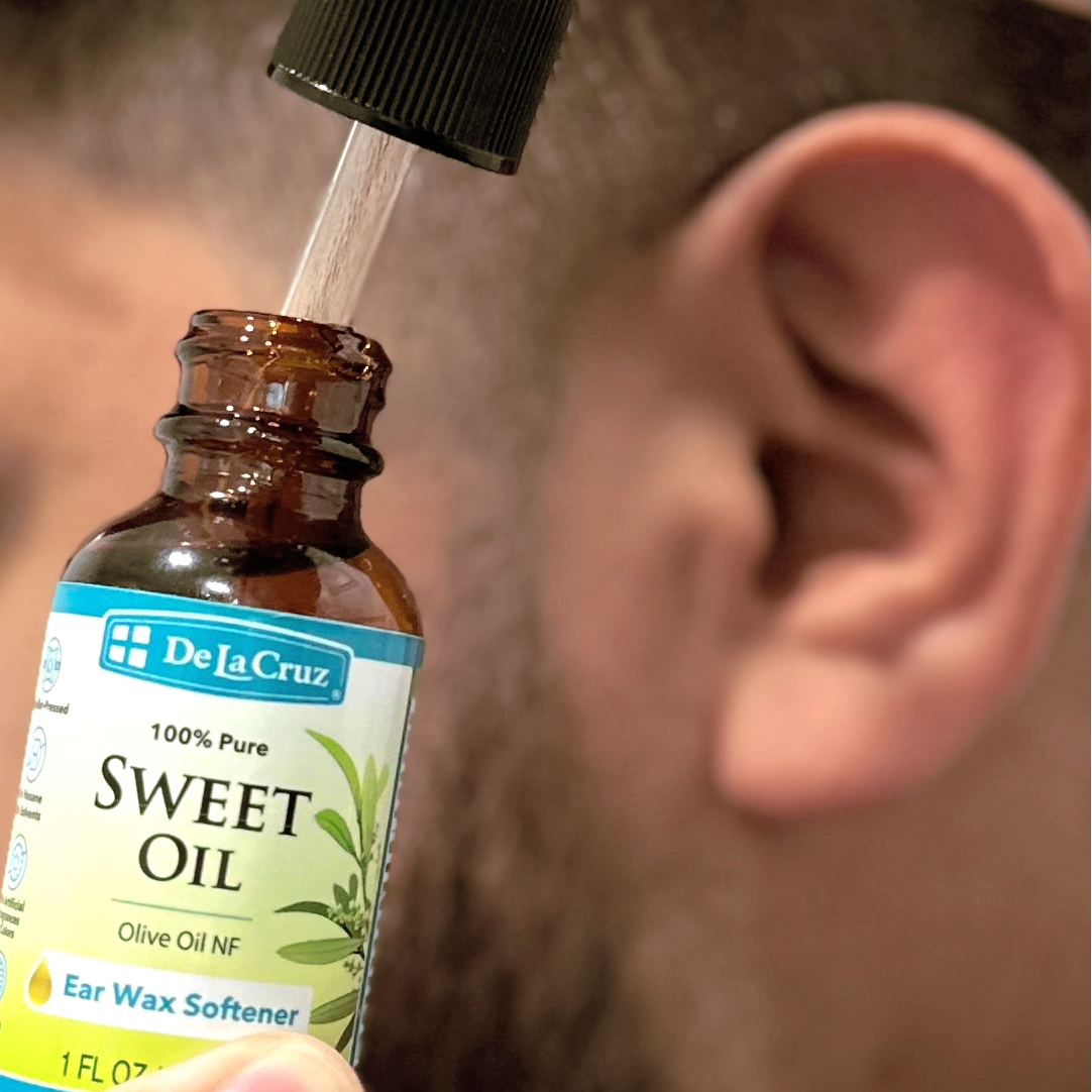 A close-up of a person holding an amber glass bottle of De La Cruz Sweet Oil Ear Wax Softener while another hand dispenses the oil into a dropper above the bottle.