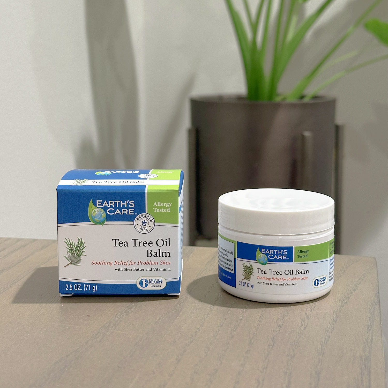 Earth's Care Tea Tree Oil Balm shown in its blue and white retail box next to an open white plastic jar with a matching label, placed on a light wood tabletop with a blurred potted plant in the background.