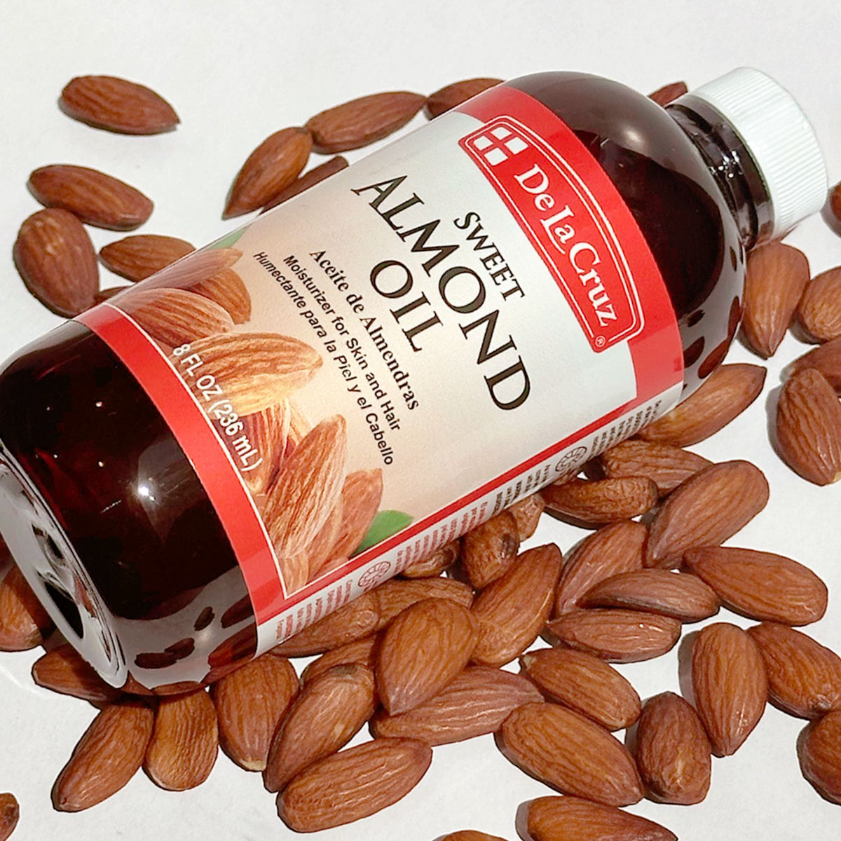 A bottle of De La Cruz Sweet Almond Oil surrounded by scattered whole almonds on a white background.