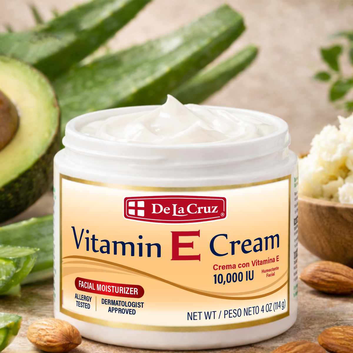 A jar of De La Cruz Vitamin E Facial Moisturizer with white cream visible at the top, surrounded by a styled flat-lay of a sliced avocado, aloe vera leaves, almonds, and a small bowl of cream on a neutral beige surface.